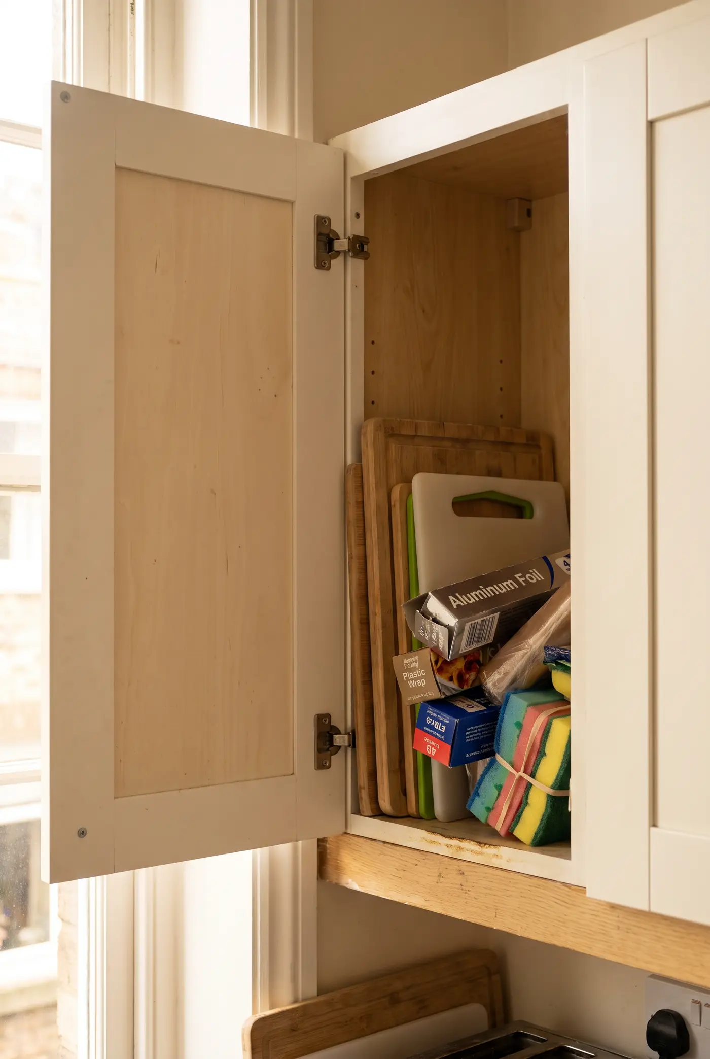 Bare inside of a kitchen cabinet door beside a shelf crammed with cutting boards and wrap boxes